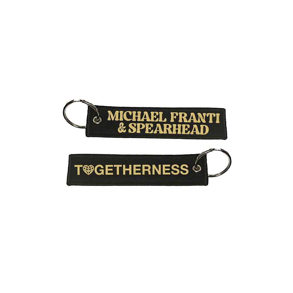 Two black fabric keychains with gold lettering, one reading ’MICHAEL FRANTI & SPEARHEAD’ and the other ’TOGETHERNESS’ with a heart symbol.