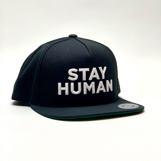 Black snapback cap with ’STAY HUMAN’ embroidered in white letters on the front.