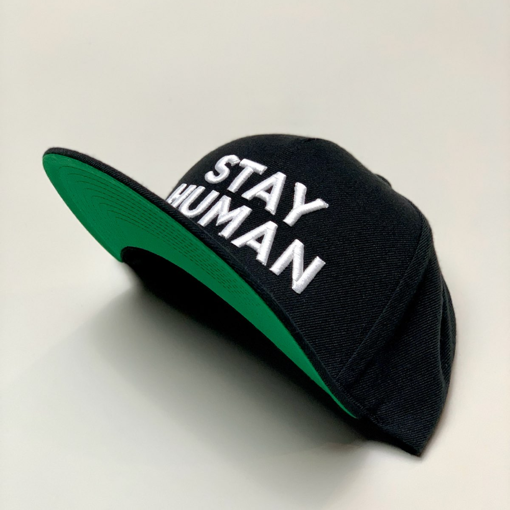 Black baseball cap with ’STAY HUMAN’ text and green underside of the brim.