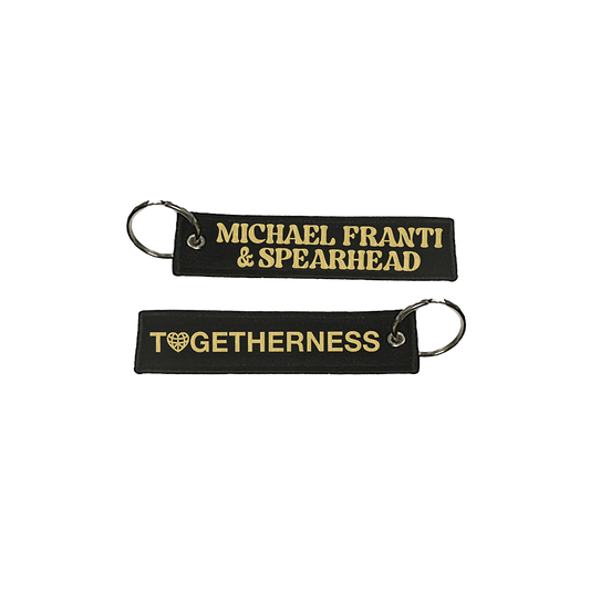 Two black fabric keychains with gold lettering, one reading ’MICHAEL FRANTI & SPEARHEAD’ and the other ’TOGETHERNESS’ with a heart symbol.